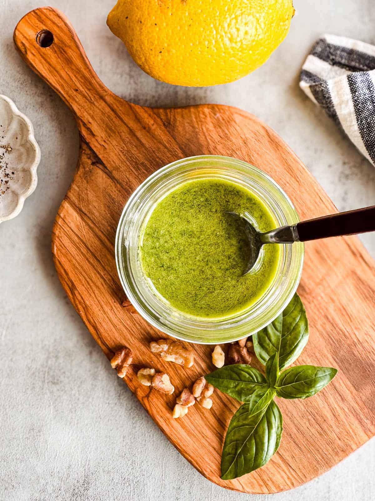 flat lay of a mason jar full of pesto on a wooden cutting board with walnuts, fresh basil, salt and lemon.