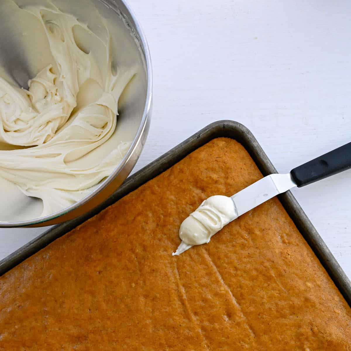 baked pumpkin bars with a bowl of cream cheese frosting and an offset spatula.