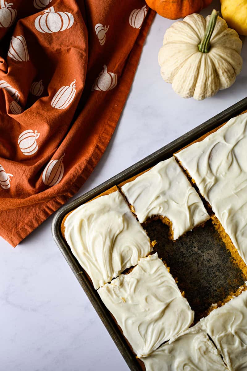 jelly roll pan with frosting pumpkin bars with one bar missing and a bite taken out of one alongside a pumpkin napkin.
