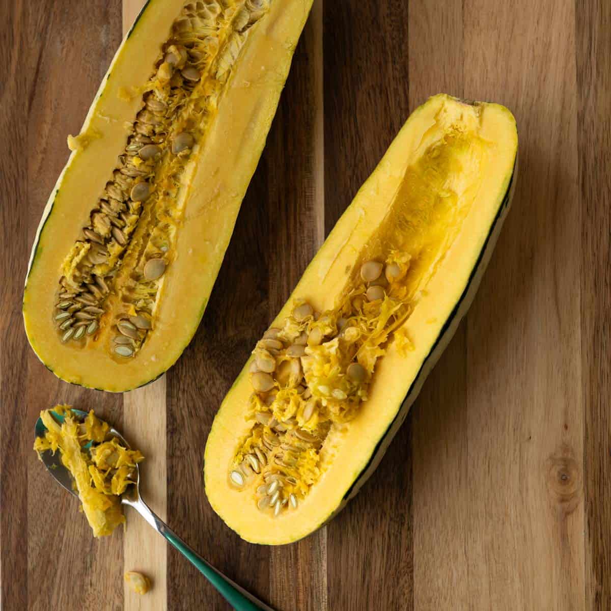 a delicata squash cut in half the long way with a spoonful of seeds on a wooden board.