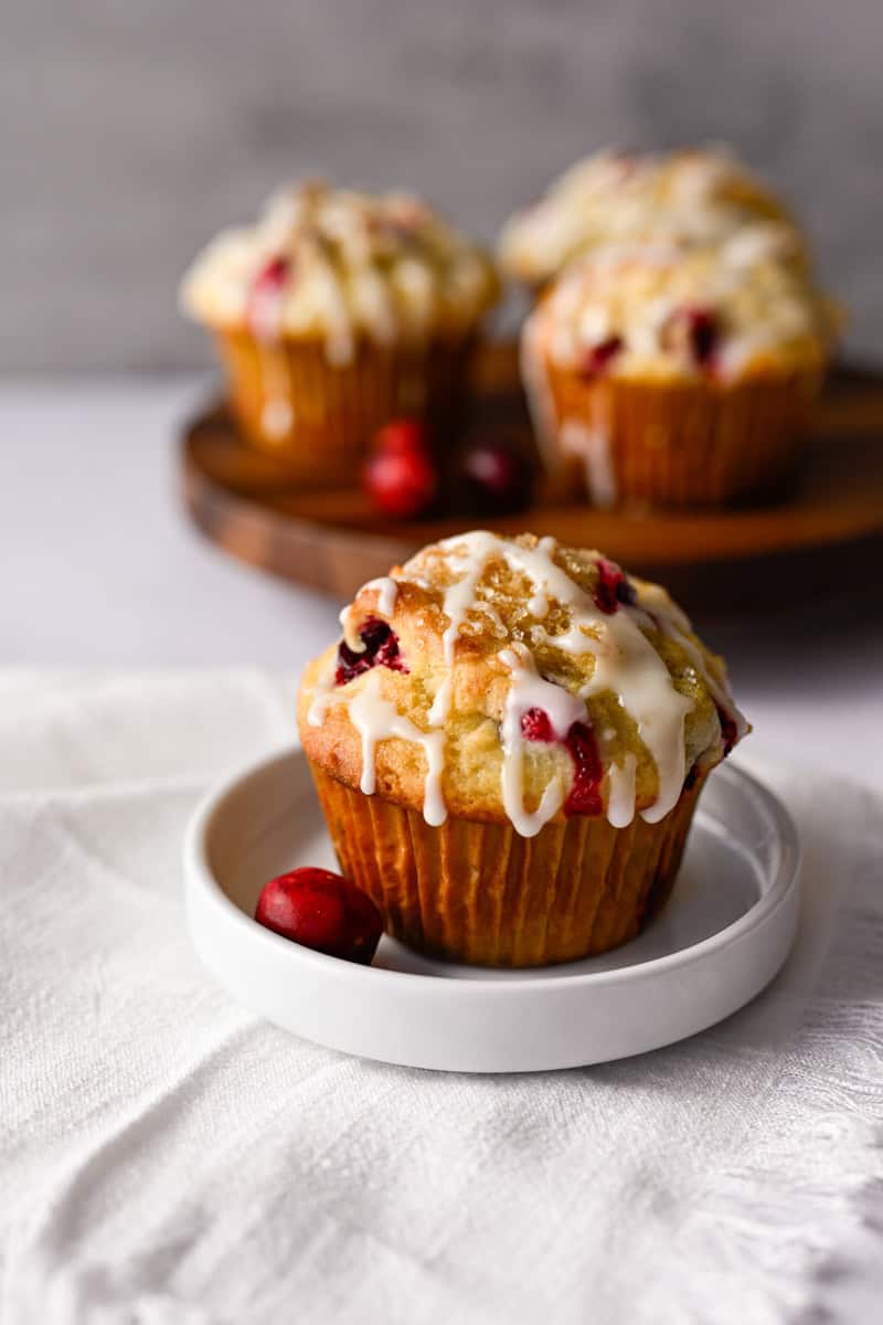 cranberry orange muffin with a glaze on a white dish with a few muffins in the background.