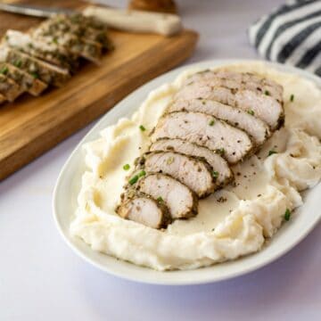 platter of mashed potatoes topped with sliced turkey tenderloin with sliced turkey tenderloin in the background on a wooden cutting board.