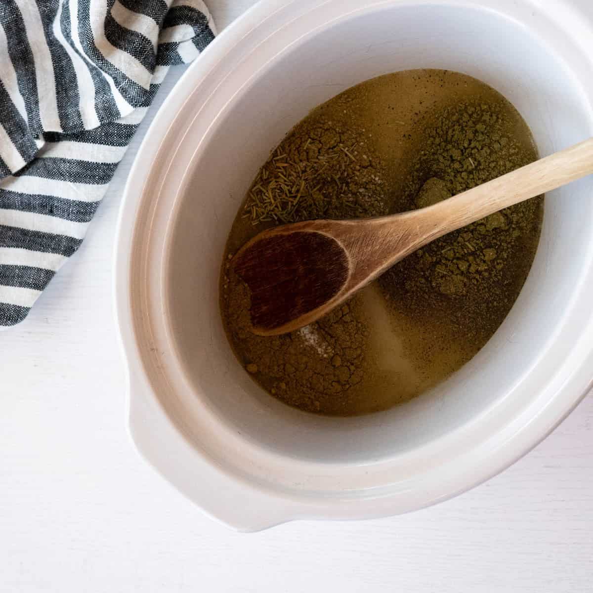 4-quart slow cooker with chicken broth and spices along with a wooden spoon.