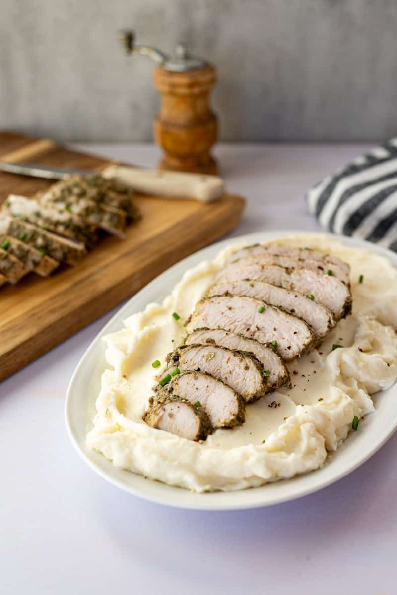 sliced turkey tenderloin on a platter of mashed potatoes with a wooden cutting board with sliced turkey in the background. 