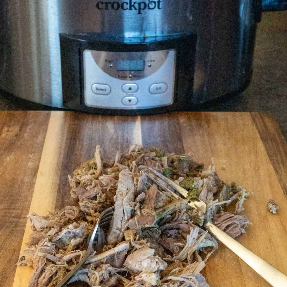 shredded pulled pork on a wooden cutting board in front of a slow cooker.
