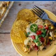 Close view of cheesy green chile chicken casserole topped with tomatoes, jalapeños, and green onions on a rustic table.