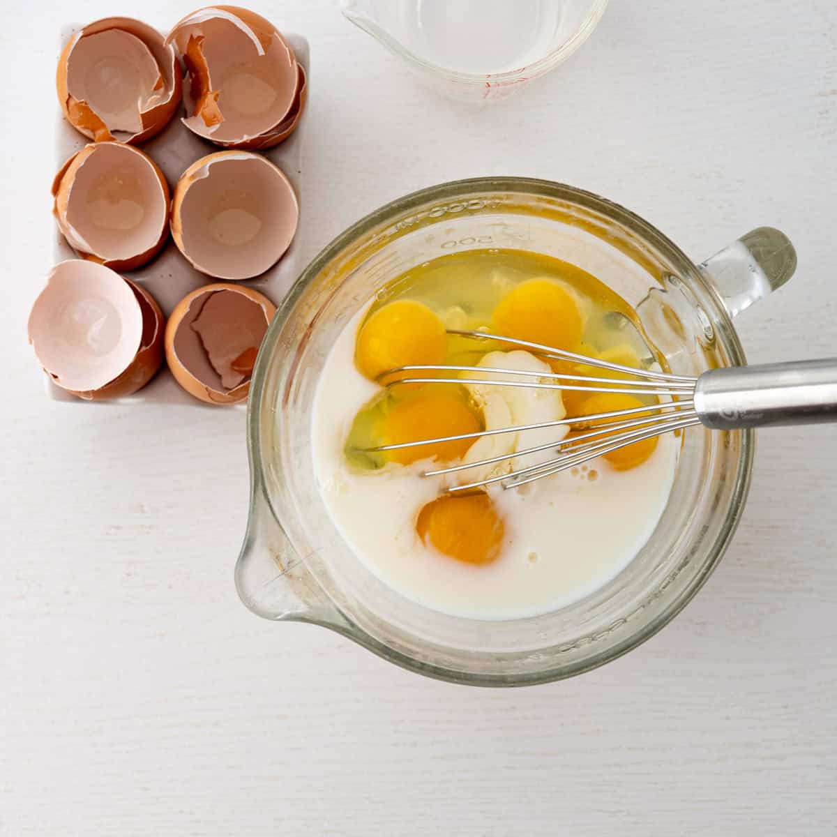 glass bowl with egg and milk with a whisk inside alongside cracked eggs.