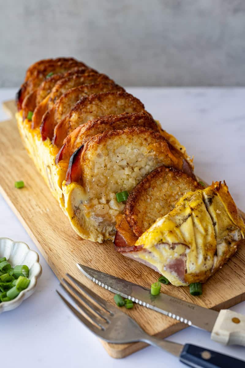sliced serving of hash brown casserole on a wooden board with a fork and knife.
