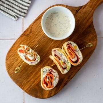 gluten free pinwheels packed with fresh veggies on a wooden cutting board alongside a small dish of ranch dressing.