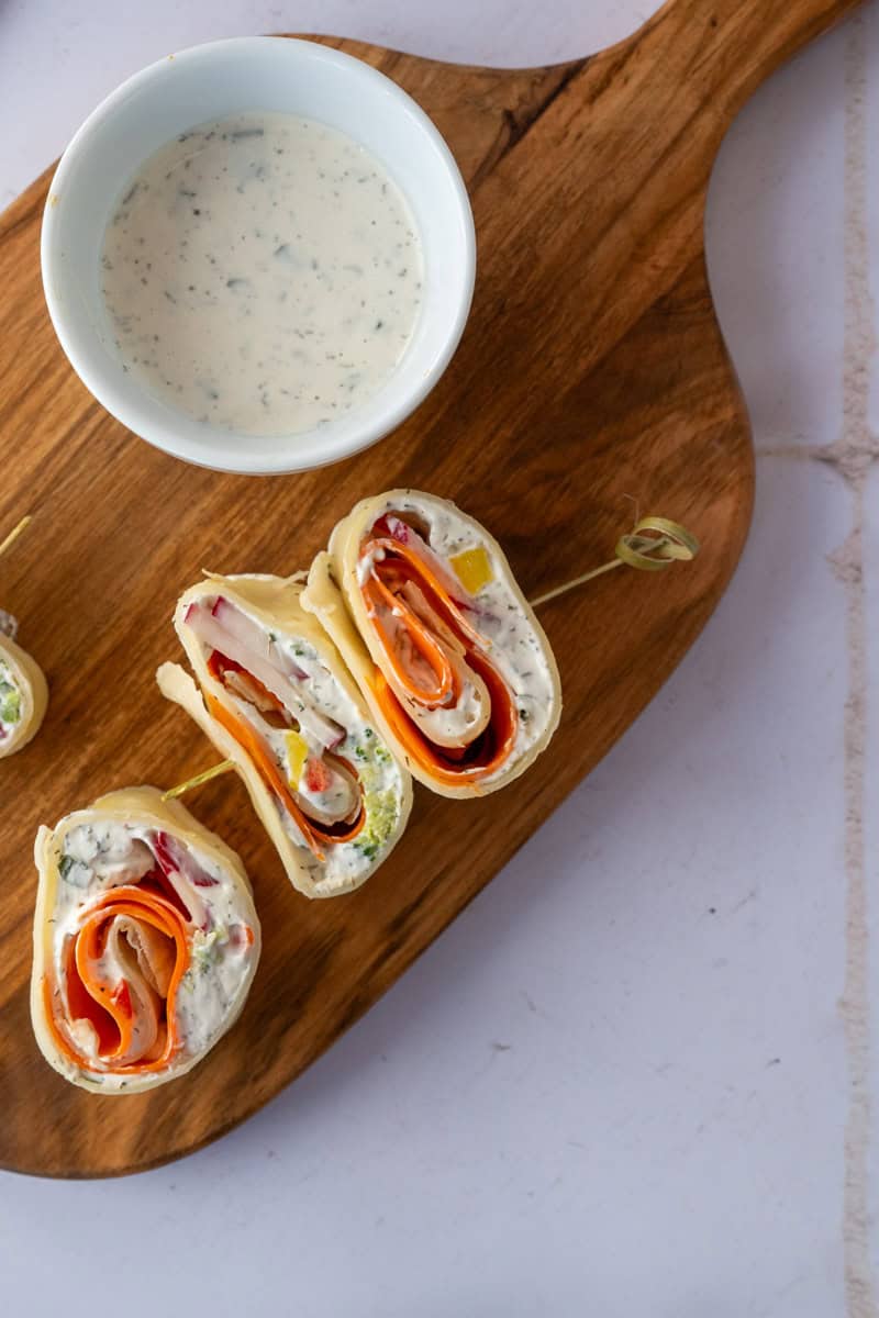 three tortilla roll ups on a wooden cutting board with a small dish of ranch dressing.