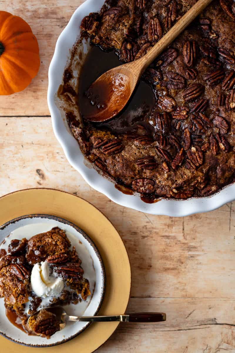 a pumpkin pudding cake in a white pie plate with a wooden spoon alongside a plate with a serving topped with ice cream.
