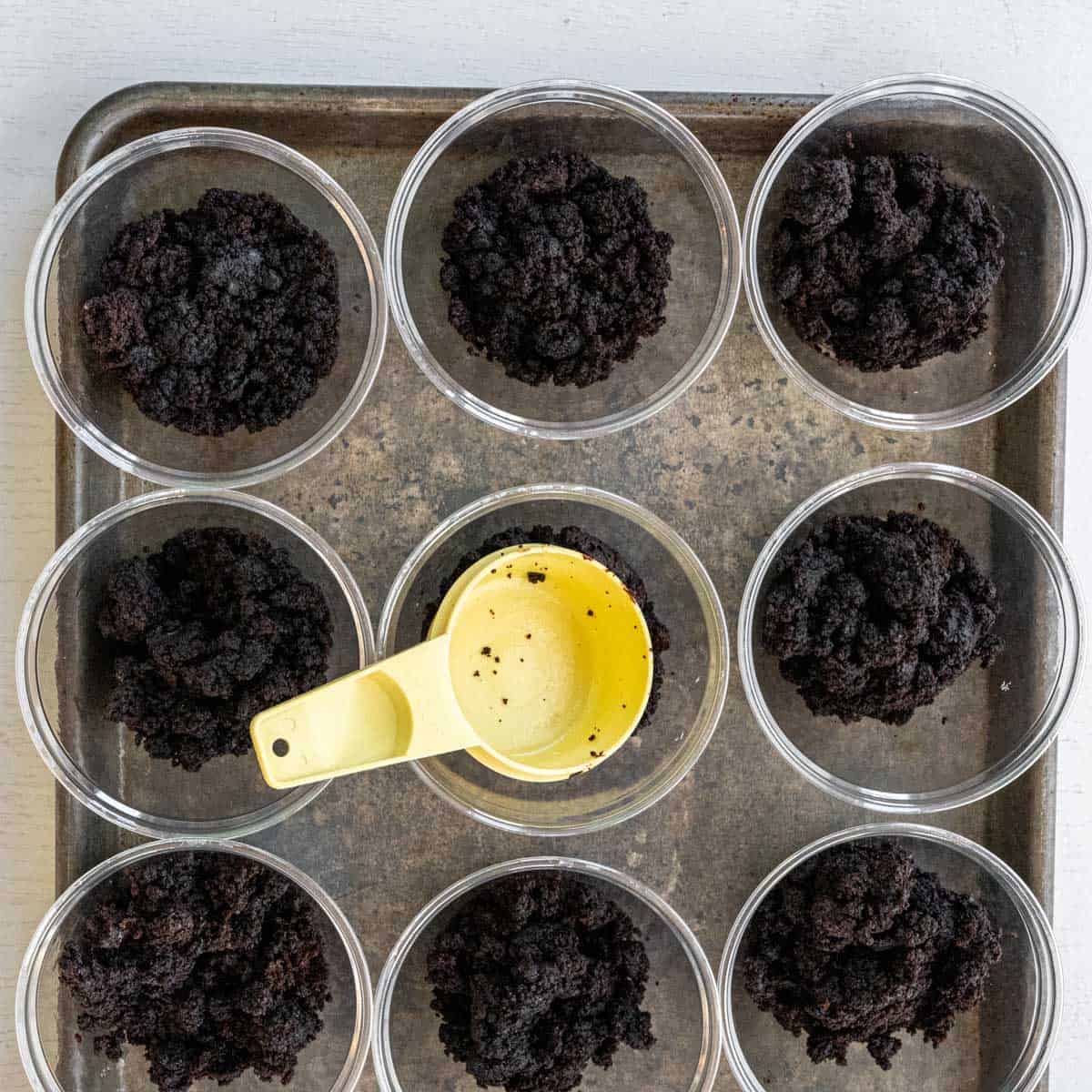 baking sheet lined with plastic cups with crumbled brownies and a yellow measuring cups.
