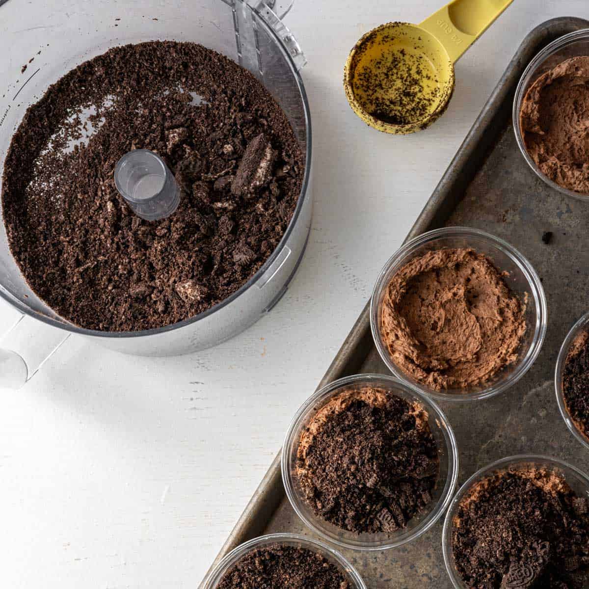 plastic cups on a baking sheet filled with brownies, chocolate pudding, and next crushed oreos alongside a food processor bowl with the remaining crushed oreos.