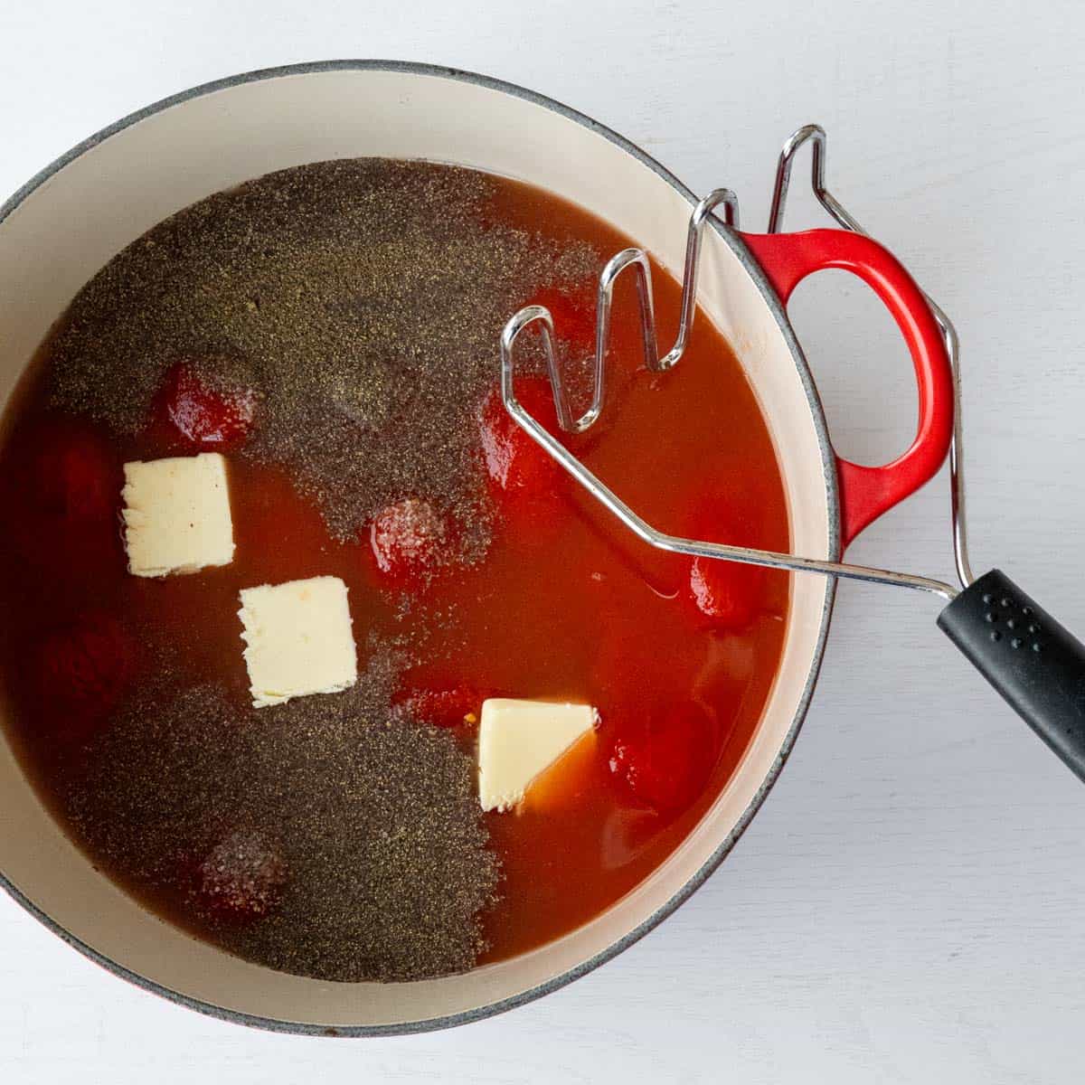 in a red dutch oven is canned whole tomatoes, spices, pats of butter and a potato masher.
