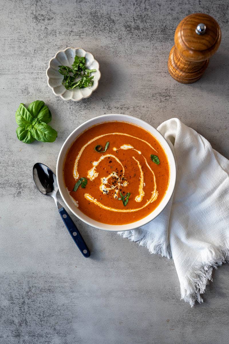 bowl of tomato soup with heavy cream swirled in alongside fresh basil and a pepper grinder.