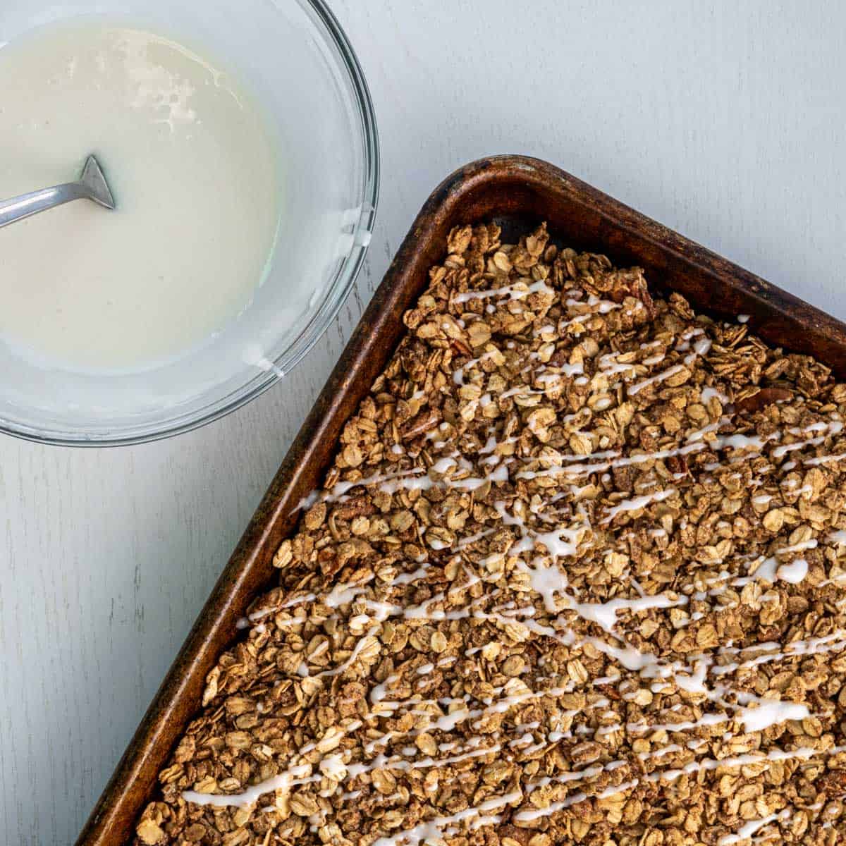 a small bowl of glaze next to pan of baked granola with drizzled glaze.
