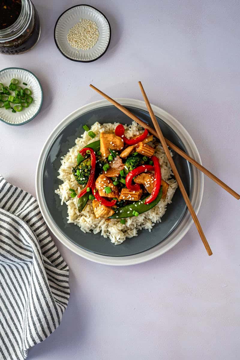 plate of chicken stir fry on a bed of white rice with chop sticks.