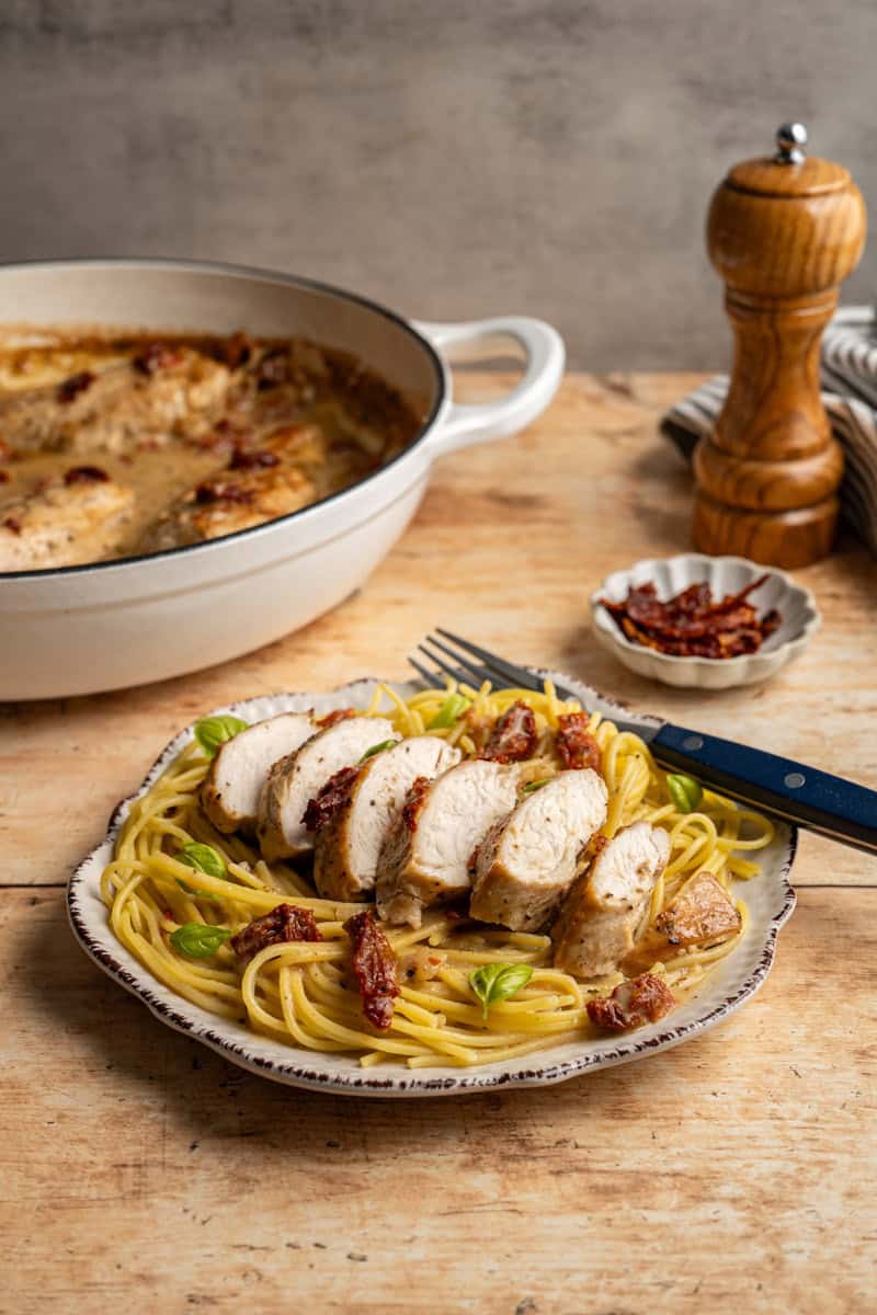 a plate of sliced marry me chicken on top of noodles, with a pan of marry me chicken in the background with a small dish of sun dried tomatoes and a pepper mill grinder.