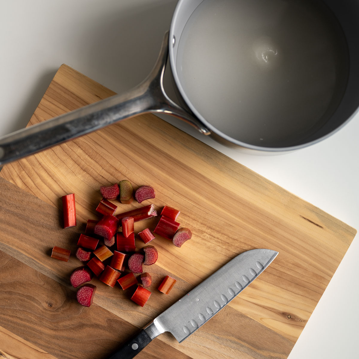 wooden cutting board with chopped rhubarb and a saucepan with sugar and water inside.