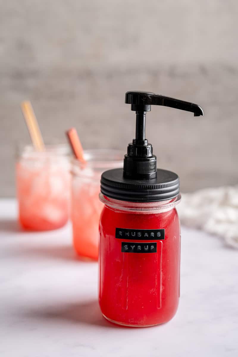 rhubarb syrup in a mason jar with a pump alongside two mason jars filled with lemonade and syrup.