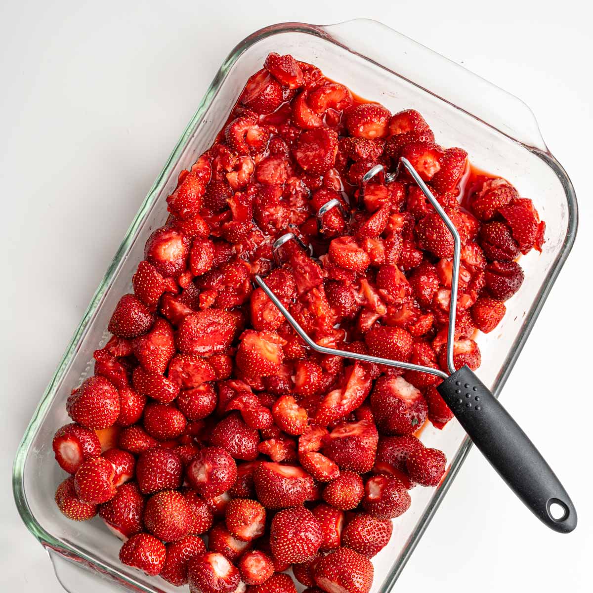 glass baking dish filled with strawberries with half mashed with a potato masher.
