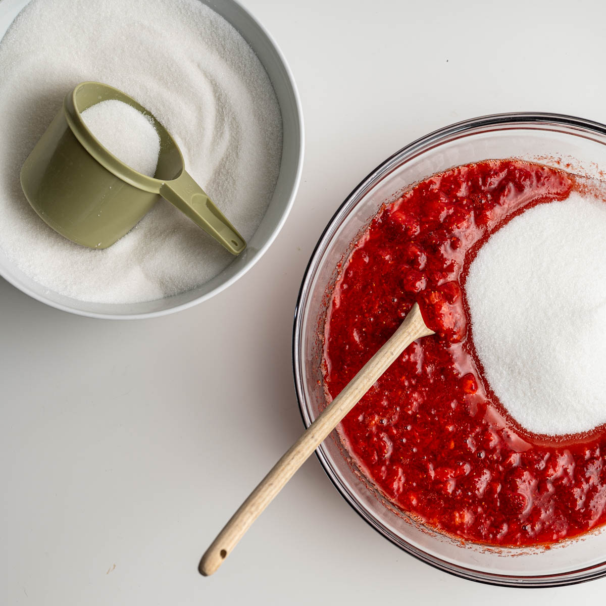 two bowls, one with sugar and a measuring cup, the other with mashed strawberries, sugar and a spoon.
