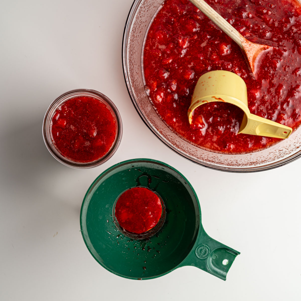 large glass bowl with stirred strawberry jam, with a mason filled and another mason jar being filled with a wide mouth funnel.