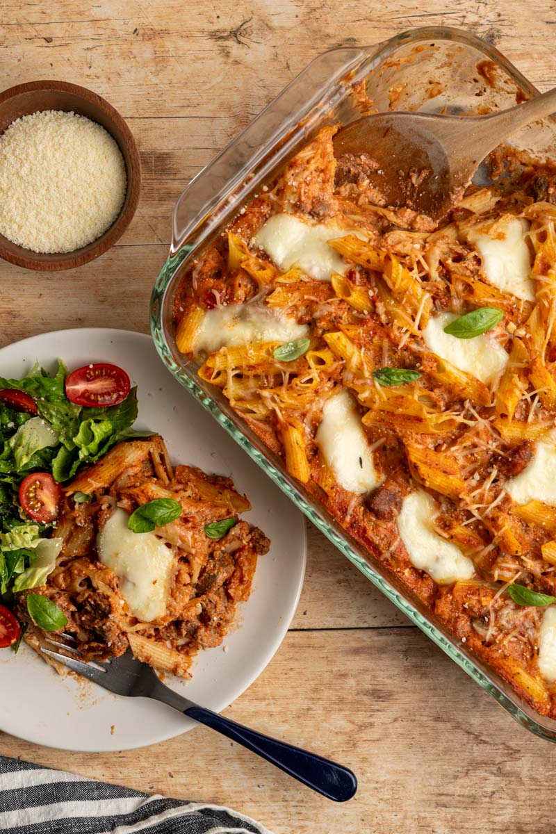 baked ziti in a casserole pan, alongside a serving on a plate with a side salad. 
