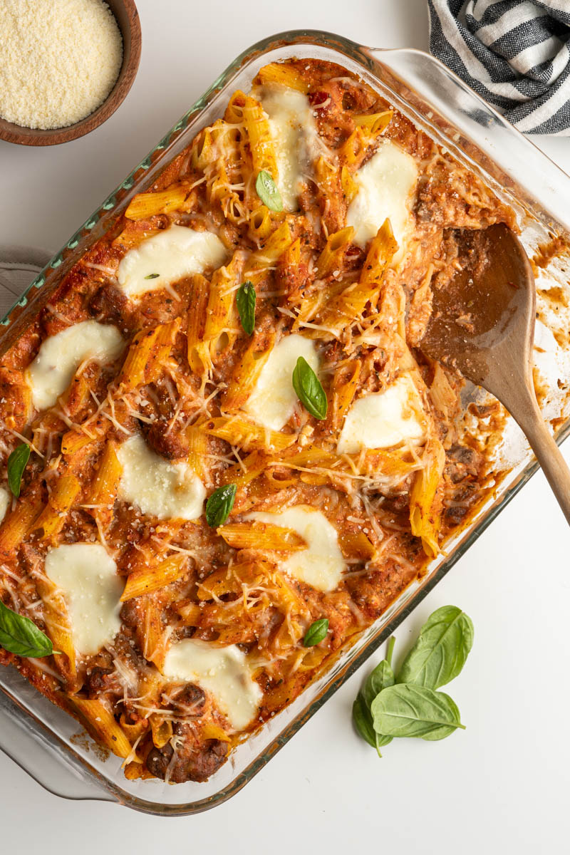 baked ziti topped with fresh basil, grated parm and a wooden spoon taking out a serving.