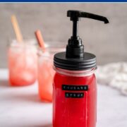 mason jar with a syrup dispenser filled with rhubarb syrup with two drinks in mason jars in the background with a rhubarb stalk as a garnish.