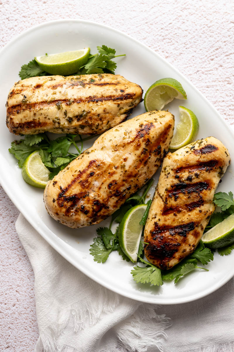 three grilled chicken breasts on a platter with quarted limes and fresh cilantro.