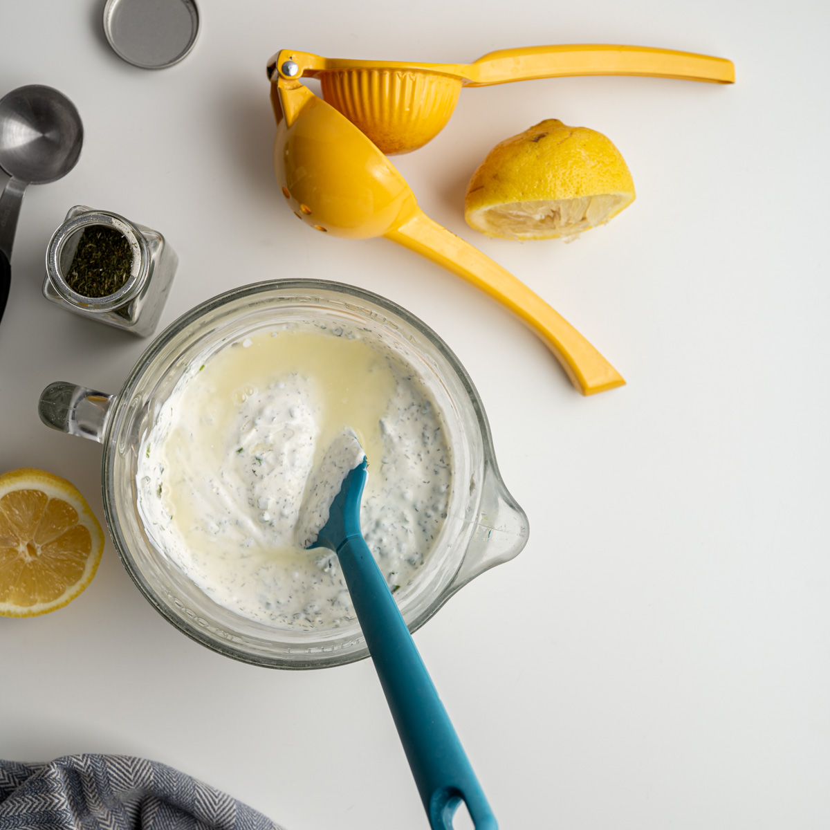glass bowl with all ingredients before stiring the vegetable dip. Alongside a citrus juicer and an opened bottle of dried dill.