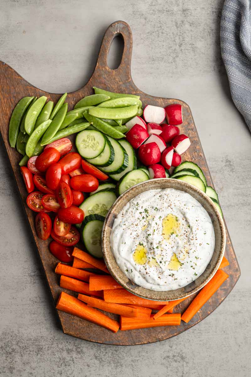 wooden board with a bowl of vegetable dip surrounded by fresh vegetables.