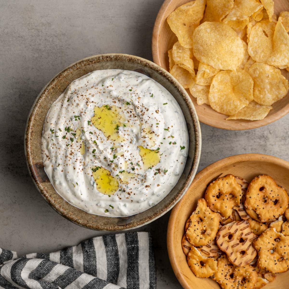 one bowl of vegetable dip alongside a bowl of potato chips and a bowl of pretzels.