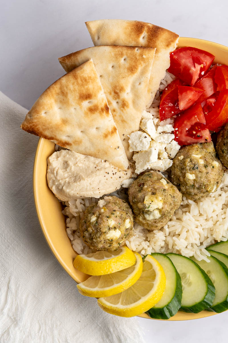 rice bowl filled with greek chicken meatballs with chopped tomatoes, sliced cucmbers, lemon slices, a spoonful of hummus and pita bread.