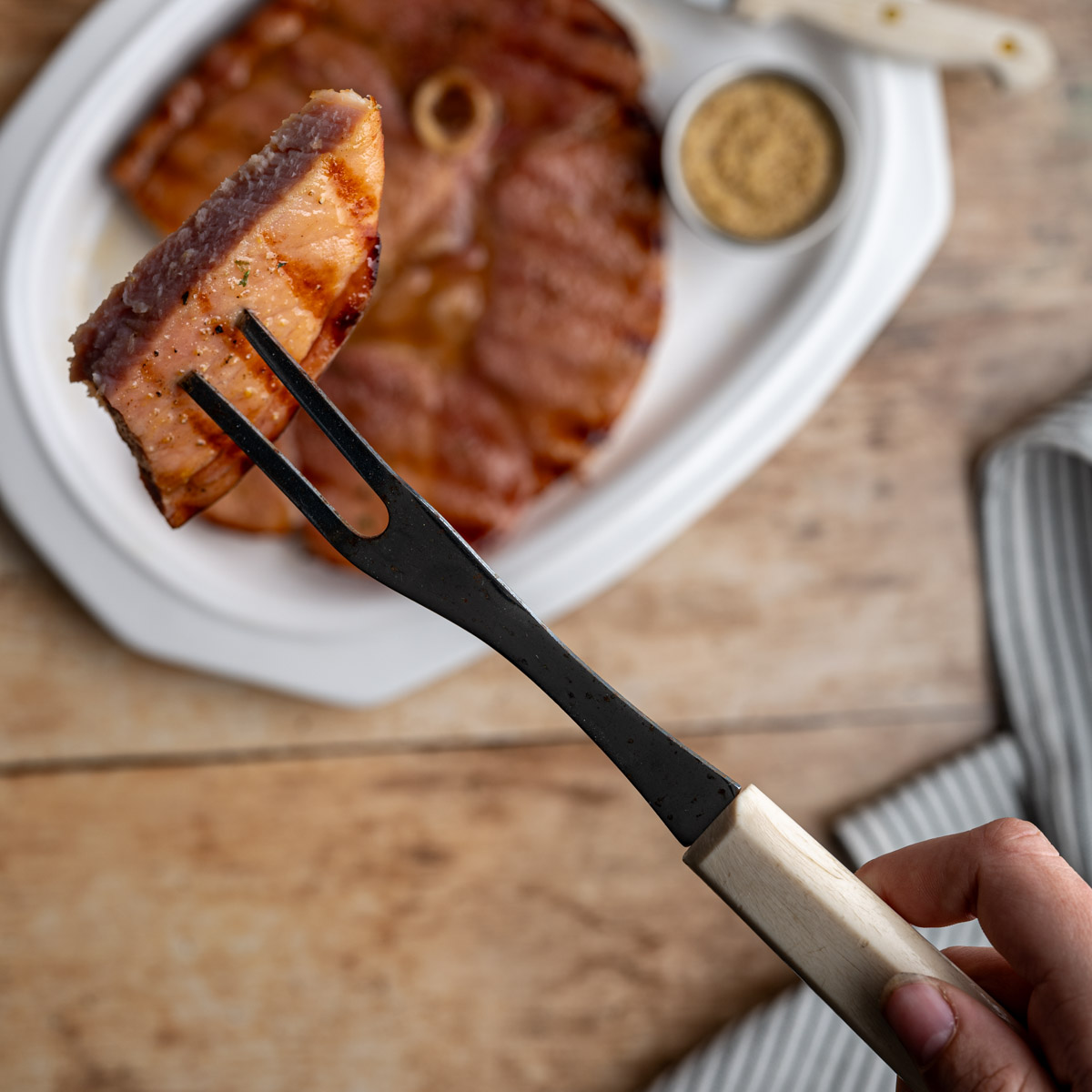 piece of grilled ham steak on a meat fork above a platter with a whole ham steak on it.