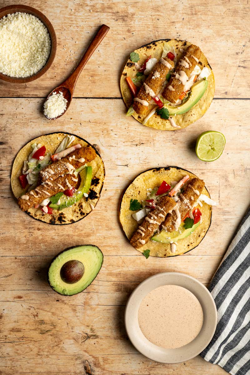 three fish stick tacos laid flat on a wooden board with a halved avocado, squeezed lime and cojita cheese.