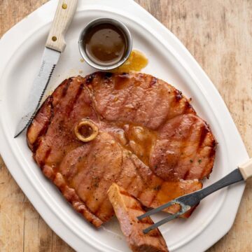 grilled ham steak on a white platter with a piece cut off with extra glaze in a small dish.