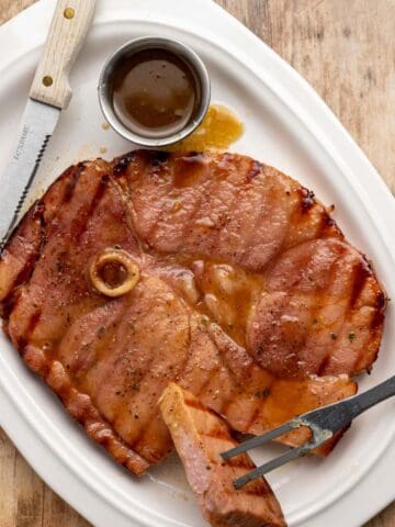 grilled ham steak on a white platter with a piece cut off with extra glaze in a small dish.