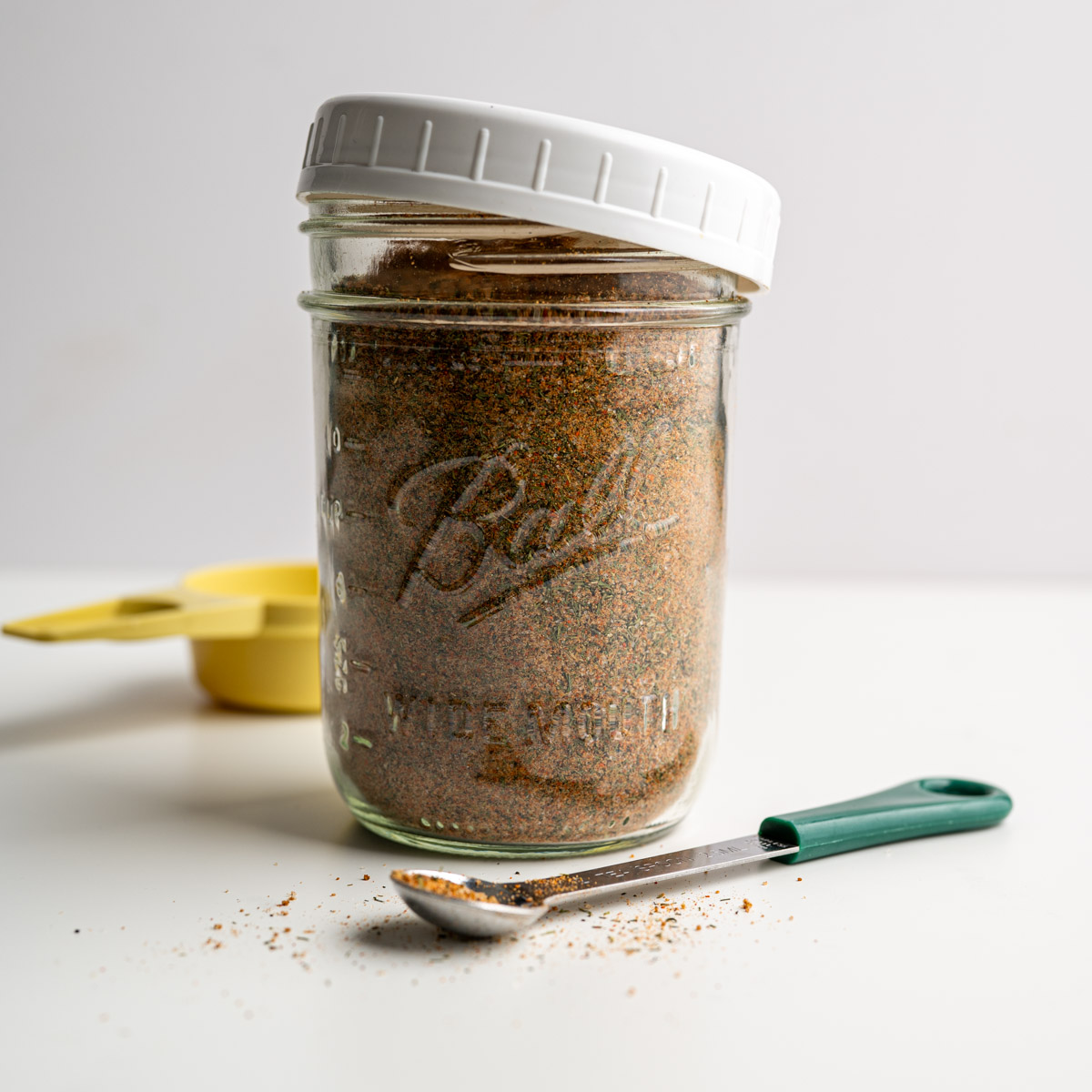 mason jar with the cover adjar filled with bloody mary seasoning with measuring cups and spoons alongside it.