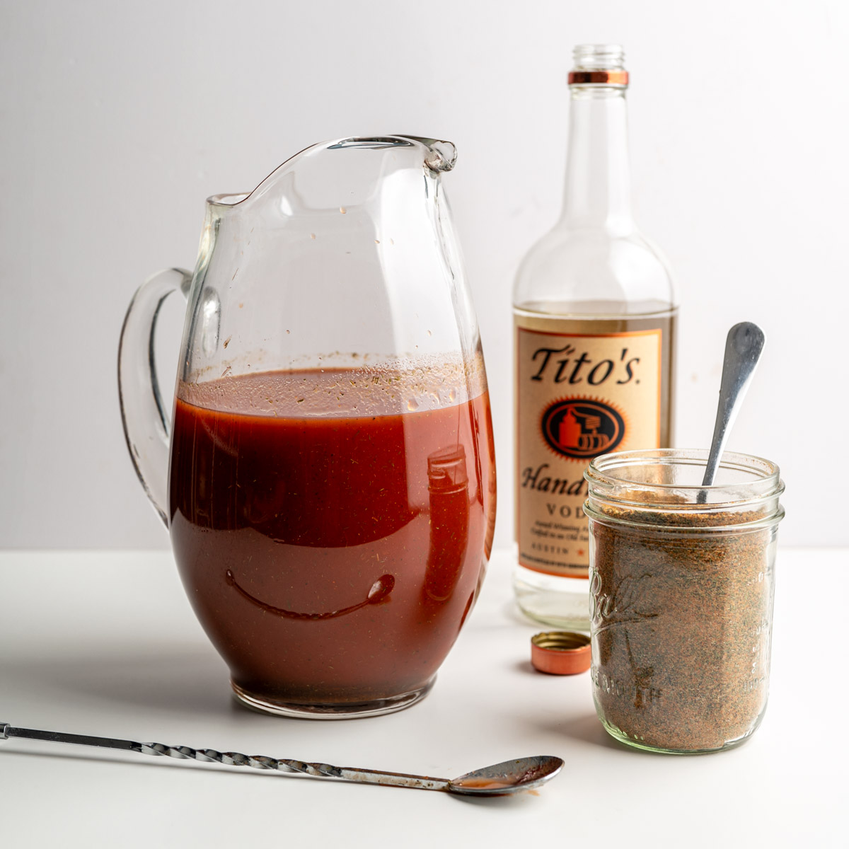pitcher filled with tomato juice next to a mason jar of bloody mary seasoning and a bottle of vodka.