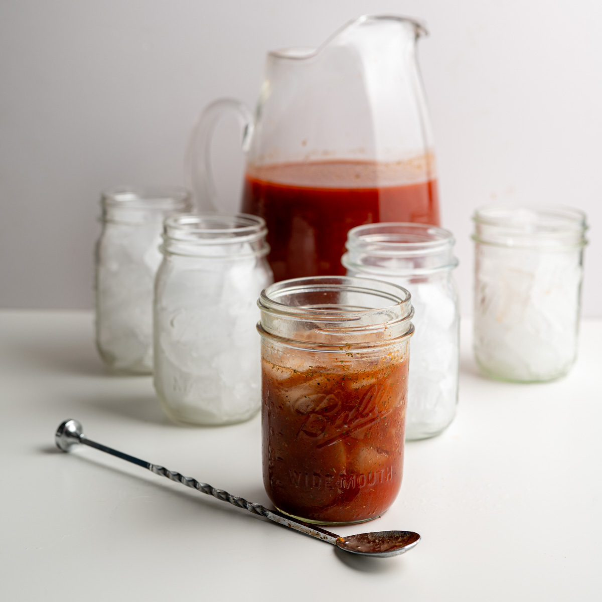 a pitcher of bloody mary mix surrounded by mason jars filled with ice.
