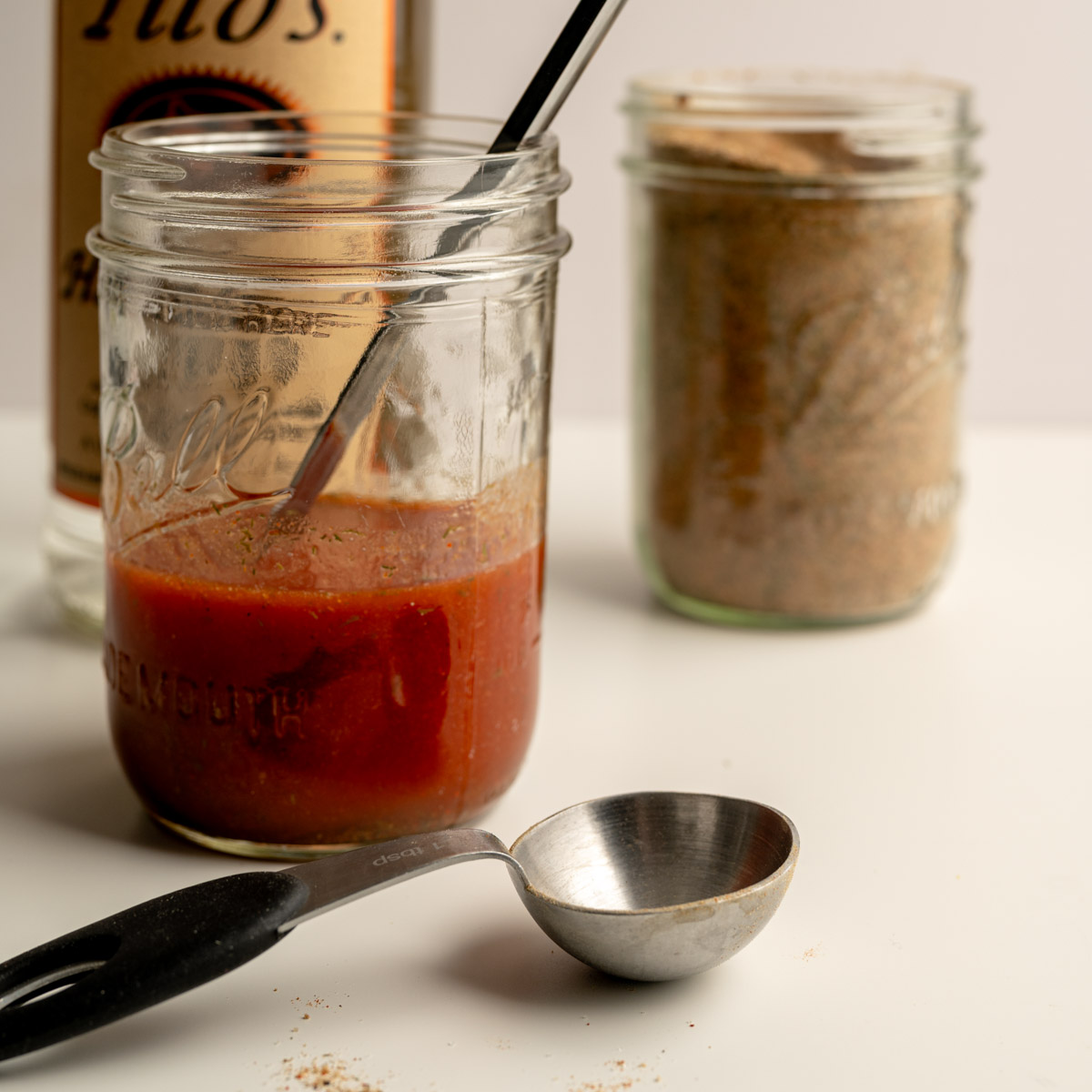 mason jar filled with tomato juice, in front of a bottle of vodka and a jar of seasoning.