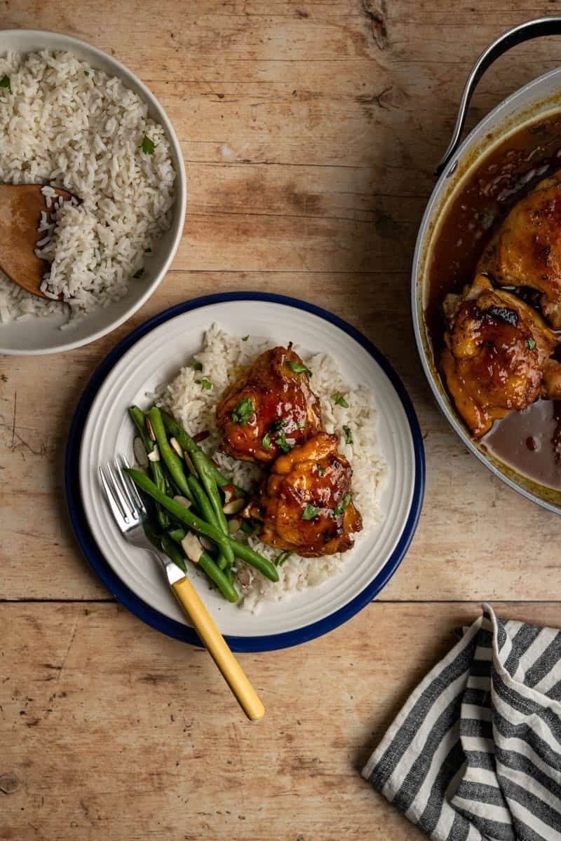 plate with apricot curry chicken thigh alongside coconut rice, green beans on a wooden table.