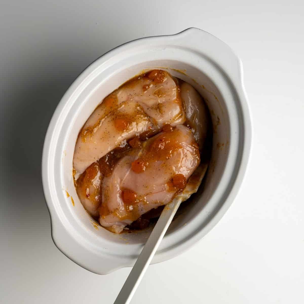 four raw chicken breasts with apricot curry sauce in a white slow cooker.