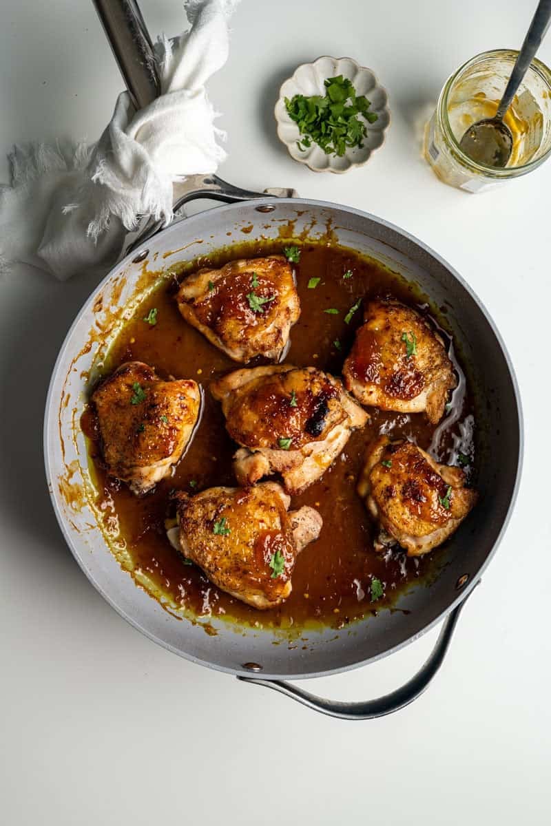 baked chicken apricot thighs in a skillet alongside chopped cilantro and an empty apricot jam jar.