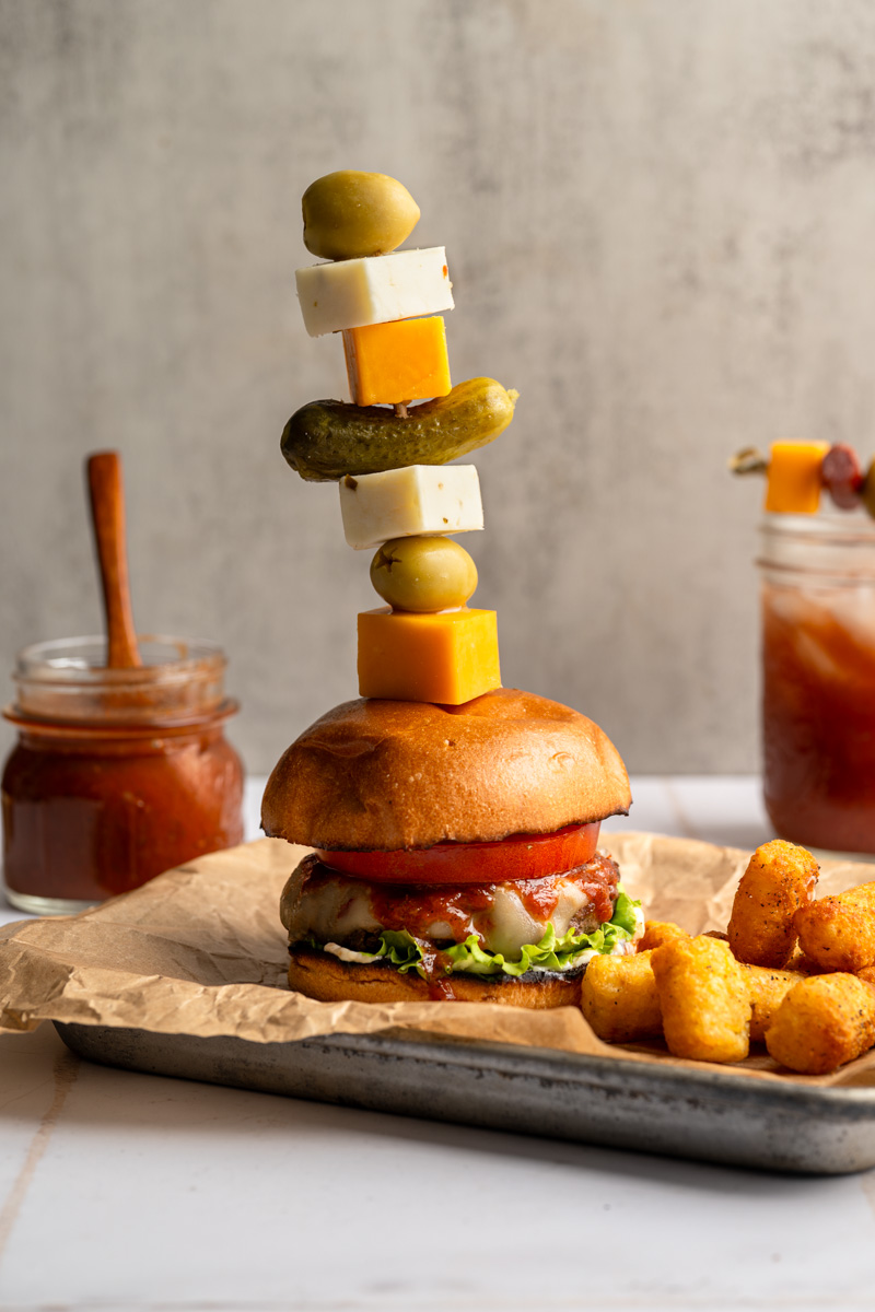 bloody mary burger with a tall skewer of garnishes, a jar of bloody mary ketchup and a bloody mary drink in the background. 