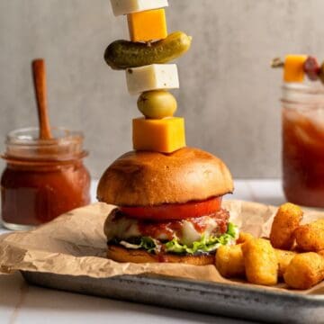 close up of bloody mary burger stacked with garnishes next to bloody mary ketchup and a bloody mary drink.