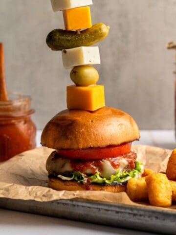 close up of bloody mary burger stacked with garnishes next to bloody mary ketchup and a bloody mary drink.