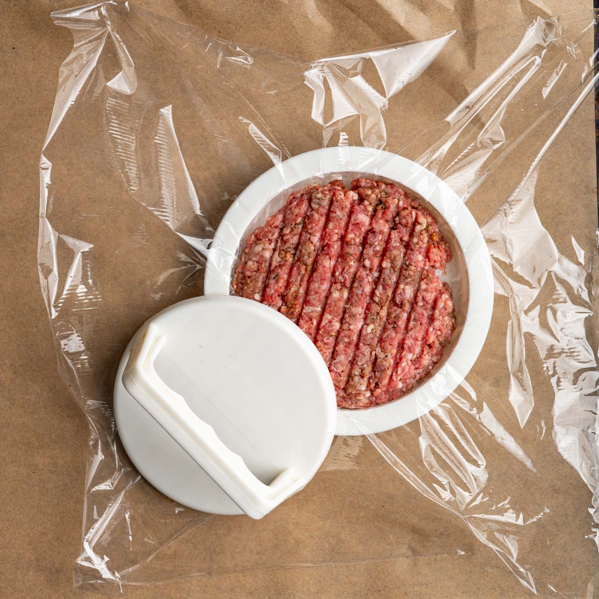 raw seasoned burger pressed into a burger press with plastic wrap on a baking sheet.